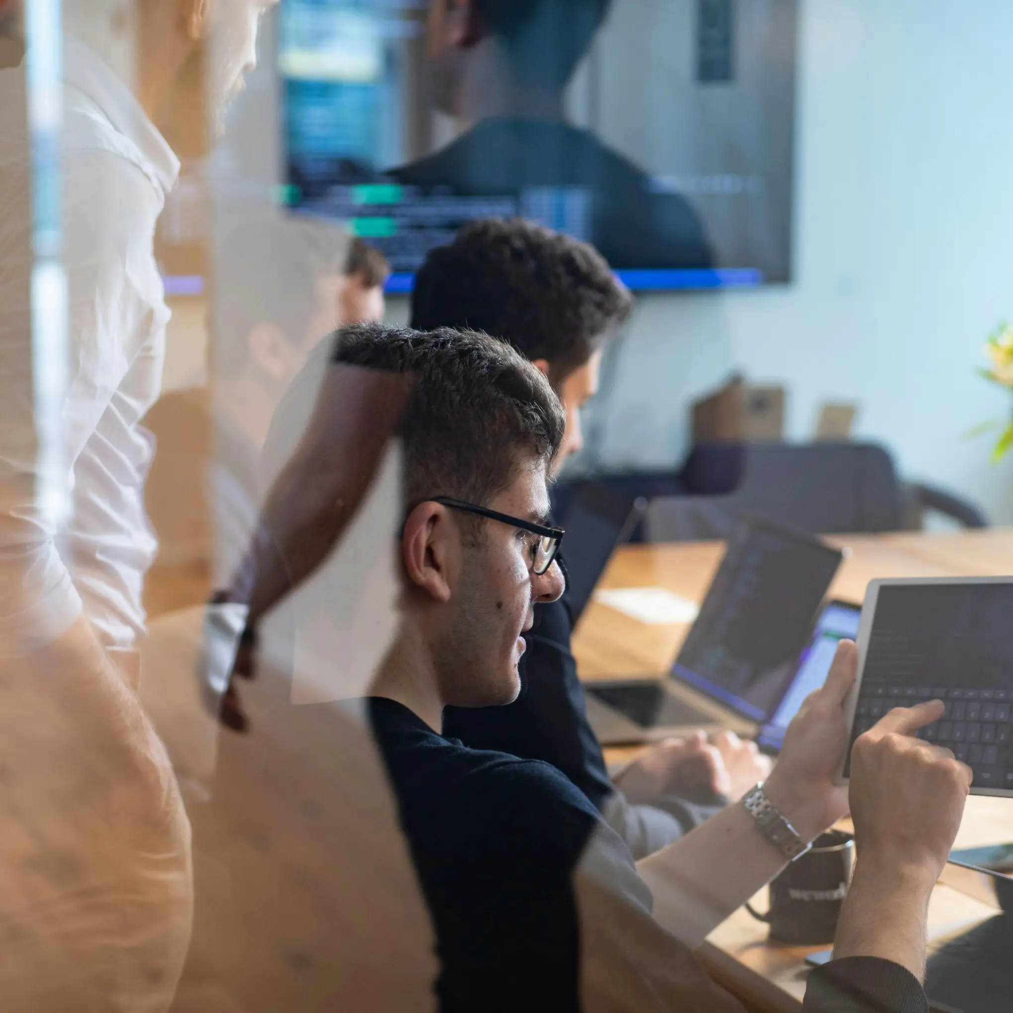 Coworkers working together at a conference table, with one person using a tablet and others using laptops in a bright office.
