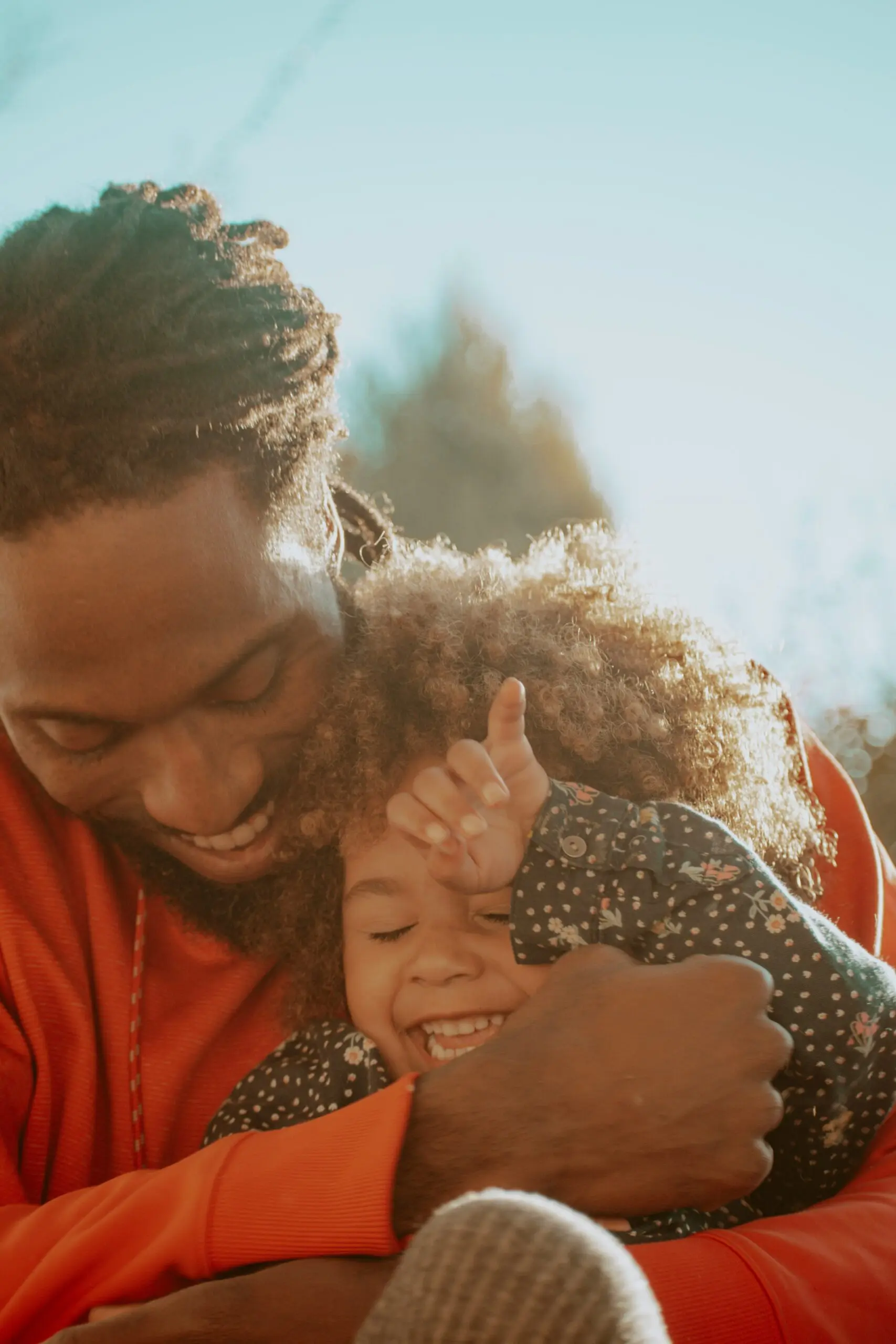 A smiling father hugs and playfully cuddles his laughing young child outdoors in warm sunlight.