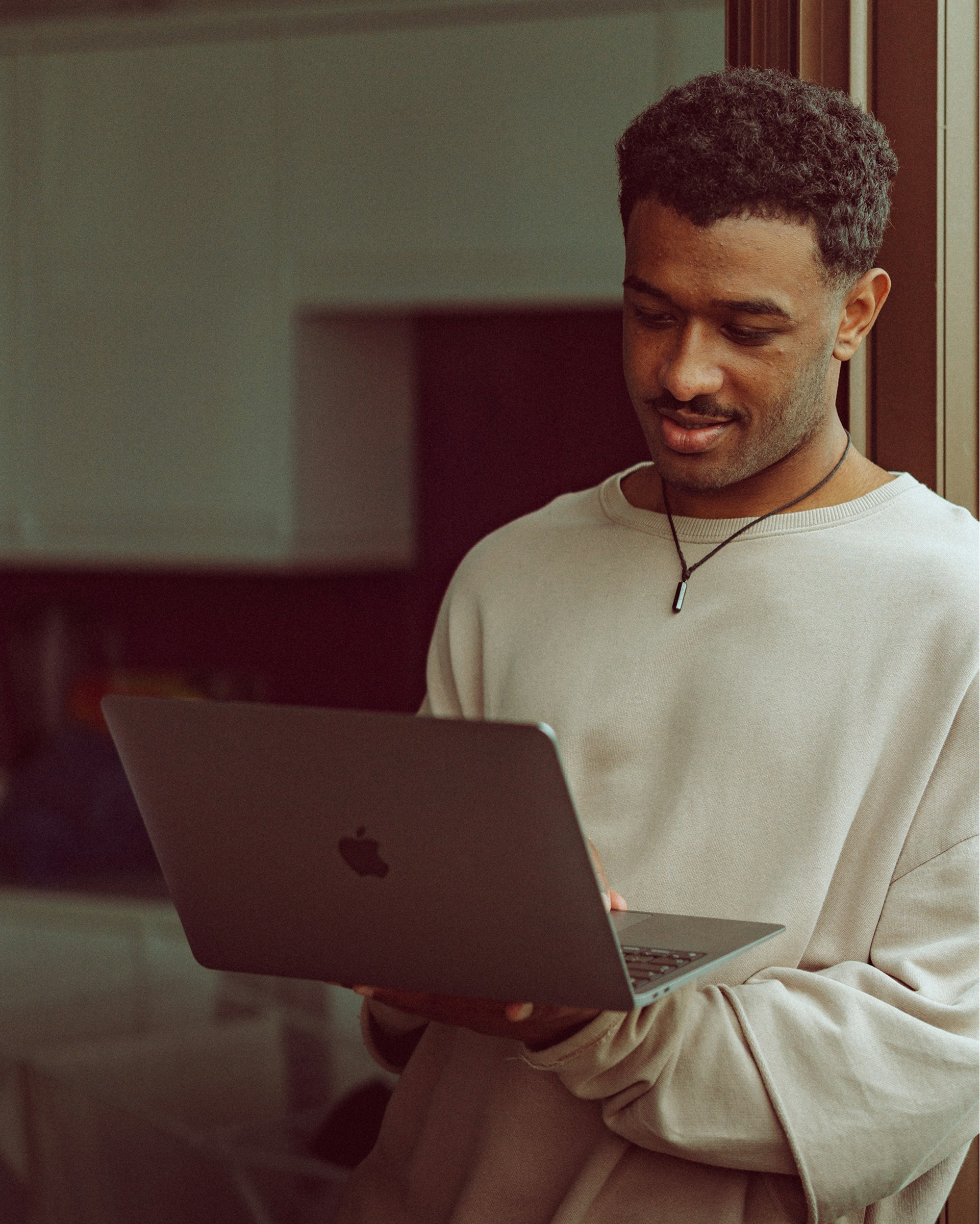 Man standing indoors and looking at an open laptop he holds in both hands.